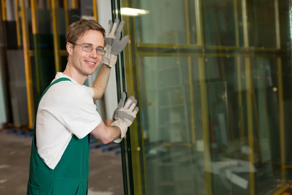 Professioneller Glaser beim Handling von Isolierglas in der Werkstatt Glaser mit Schutzkleidung hebt eine große Glasscheibe in einer Werkhalle mit Handschuhe